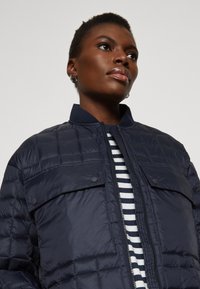 Marineblå quilted puffer-jakke med stålkrage, frontlukkning med lynlås og trykknap-lommer. Har en stripet skjorte under.