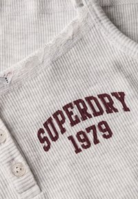 Lichtgrijze, geribbelde tanktop met kantafwerking en bordeauxrode "SUPERDRY 1979" opdruk. Opvallende knoopdetails aan de linker voorkant.