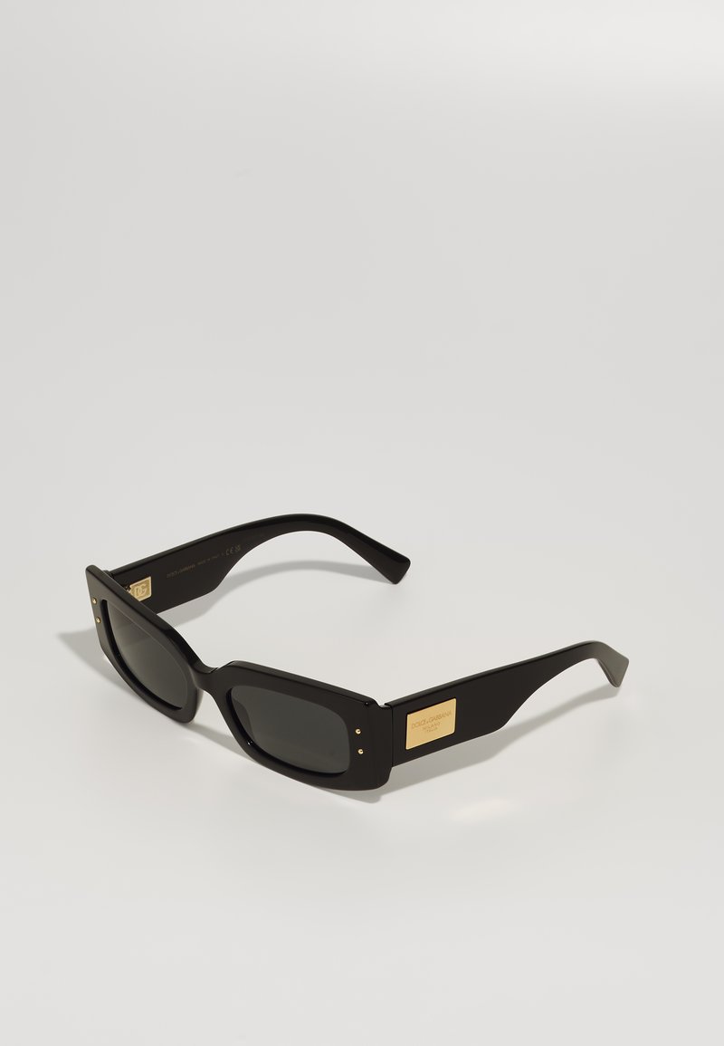 Óculos de sol retangulares pretos com lentes escuras, armação de plástico espesso e detalhe em logotipo dourado na lateral. Textura suave com bordas arredondadas.