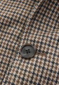 Ruskea ja beige houndstooth-kangas, jossa on sileä pinta, sekä kiiltävä musta nappi, jossa on neljä reikää, ommeltu reunaan.