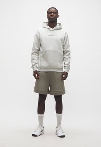 Άνδρας στέκεται φορώντας ανοιχτό γκρι φούτερ με κουκούλα, χακί σορτς, λευκές κάλτσες και λευκά παπούτσια Nike σε απλό λευκό φόντο.