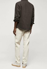 Man die met zijn rug naar voren staat en een opgerolde donkerbruin shirt, lichtbeige spijkerbroek en lichtbeige sneakers draagt, staat voor een effen achtergrond.