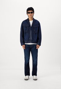Jachetă din denim cu închidere prin buton, două buzunare la piept și mâneci lungi. Asociată cu blugi închiși la culoare și pantofi albi. Accesoriile includ ochelari de soare închiși.