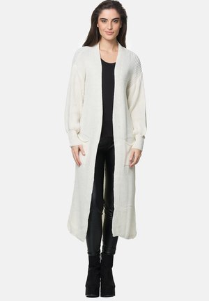 Lang, open voorkant cardigan in crème, gemaakt van een textuur gebreid materiaal. Heeft drop schouders en twee voorzakken. Gecombineerd met een zwarte top.