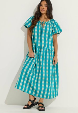 Femme aux longs cheveux foncés portant une robe midi à carreaux turquoise et blancs avec des manches bouffantes et des sandales noires à boucle, debout contre un mur blanc.