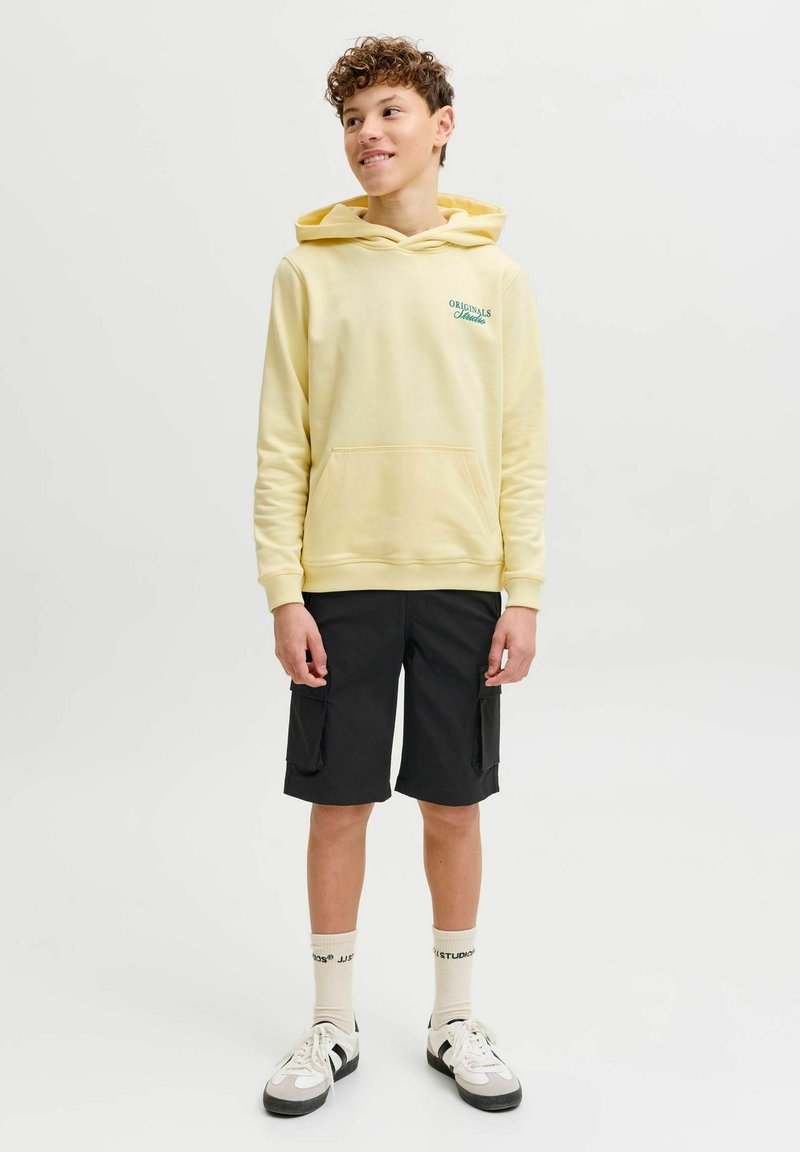 Adolescent debout, portant un sweat à capuche jaune pâle, un short cargo noir, des chaussettes blanches de marque et des baskets blanches, regardant à droite et souriant.