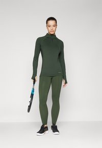 Zöld sportfelső félcipzárral, zöld leggingsszel párosítva. Fekete sportcipő, kék teniszütőt tartva.
