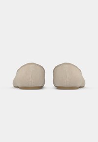 Chaussures slip-on en tissu beige avec une texture côtelée, un bout arrondi et une semelle plate. La vue de dos montre un design sans couture sans ornements.