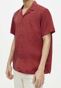 Chemise à manches courtes, boutonnée, de couleur rouge foncé avec une poche poitrine unique, en tissu léger. Présente un col classique et une coupe décontractée.