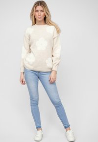 Helles beiges Strickpullover mit großen weißen Blumenmustern, Rundhalsausschnitt und gerippten Bündchen, kombiniert mit hellblauen Skinny Jeans und weißen Sneakers.
