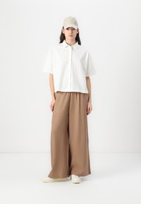 Camisa blanca texturizada cortada, pantalones beige de pierna ancha y una gorra clara, combinados con zapatos blancos, sobre un fondo neutro.