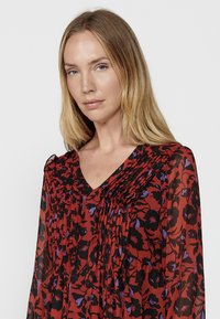 Bluză florală roșie și neagră cu decolteu în V, față plisată și mâneci lungi. Prezintă accente mov în modelul floral.