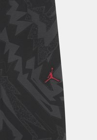 Czarne spodnie sportowe z wyrazistym szarym wzorem w zygzak. Po lewej stronie przeszyte czerwone logo Jumpman. Materiał wygląda na gładki.