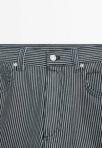 Gros plan sur un jean en denim rayé verticalement en noir et blanc, avec un bouton en métal et des passants pour ceinture à la taille.