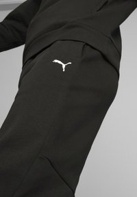 Pantalones deportivos negros hechos de tela suave, con un corte ajustado, bolsillos laterales y un logo de Puma blanco en el muslo izquierdo.