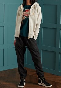 Felpa grigia chiara con zip e cappuccio, t-shirt blu navy con stampa grafica, pantaloni cargo neri e sneaker alte nere.