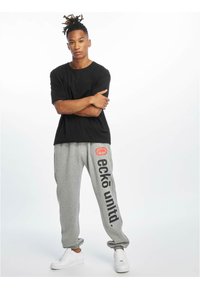 Zwart T-shirt met korte mouwen gecombineerd met grijze trainingsbroek met een groot "eckō un1t3d" logo aan de zijkant en elastische boorden, gedragen met witte sneakers.