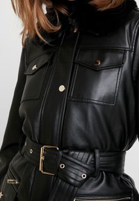 Chaqueta negra de cuero sintético con cuello de piel, herrajes dorados, dos bolsillos en el pecho y cintura con cinturón y detalles texturizados.