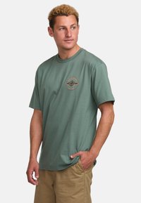Grünes Baumwoll-T-Shirt mit kurzen Ärmeln und Rundhalsausschnitt, mit einem runden Berggrafik in Braun und Orange auf der linken Brust.