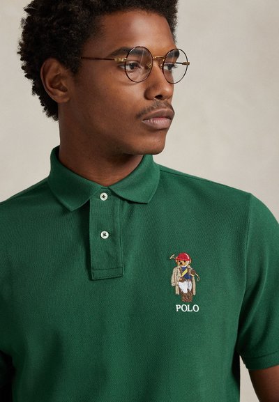 Zelena polo majica iz teksturirane tkanine, z zapenjanjem na tri gumba in vezenim logotipom medveda s rdečo kapo na levi strani prsnega koša.