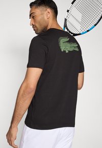 Czarny bawełniany T-shirt z zielonym motywem krokodyla na plecach. Materiał jest gładki, z krótkimi rękawami i klasycznym krojem.