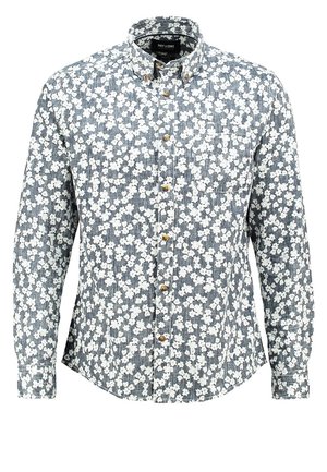 Mann trägt ein eng anliegendes schwarzes und weißes Blumenhemd mit hochgekrempelten Ärmeln, hellblaue Skinny-Jeans und schwarze Sneaker mit weißen Sohlen.