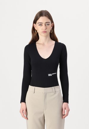 Black long-sleeve bodysuit with a deep V-neckline and a white logo on the left hip. Smooth fabric with a fitted design, paired with beige trousers.