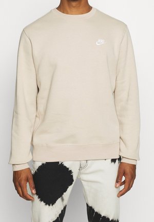 Hombre con sudadera beige de Nike y pantalones blancos con grandes parches tie-dye negros, de pie frente a un fondo liso.
