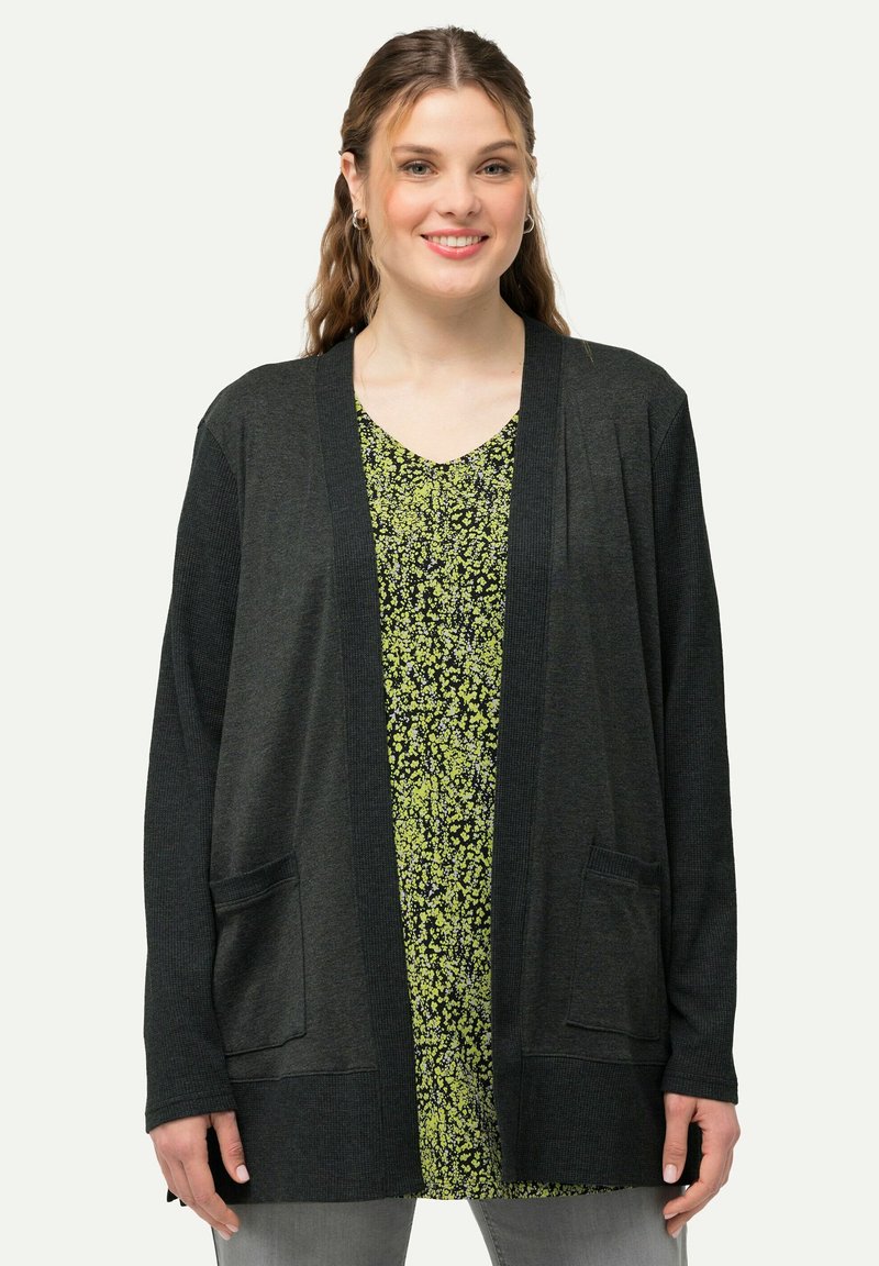 Mörkgrå cardigan med avslappnad passform, ribbad textur, öppen front och två framfickor, lagd över en blommig grön klänning.