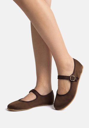 Piernas con zapatos planos estilo Mary Jane marrones con correas de hebilla sobre un fondo claro.