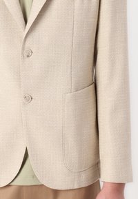 Blazer beige en tissu texturé avec deux boutons et une poche. Il présente un col cranté et des manches droites.