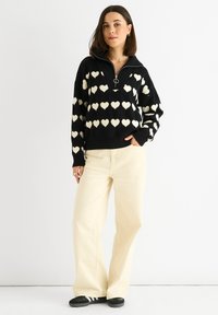 Zwarte trui met witte hartpatronen, een ritssluiting bij de kraag en een losse fit; gecombineerd met lichtcrème wijde broek en zwarte schoenen.