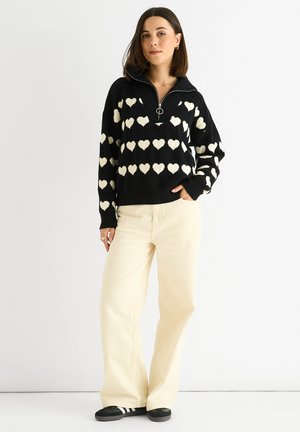 Schwarzer Pullover mit weißen Herzmustern, einem Reißverschlusskragen und lockerer Passform; kombiniert mit hellbeigen, weit geschnittenen Hosen und schwarzen Schuhen.