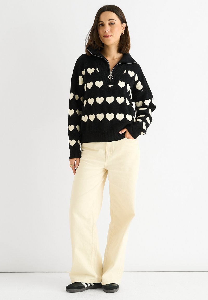 Zwarte trui met witte hartpatronen, een ritssluiting bij de kraag en een losse fit; gecombineerd met lichtcrème wijde broek en zwarte schoenen.