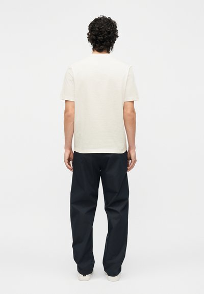 Άνδρας με σγουρά μαλλιά που φοράει ένα απλό λευκό κοντομάνικο μπλουζάκι και φαρδιά μαύρη παντελόνα, στέκεται γυρισμένος με την πλάτη προς το λευκό φόντο.