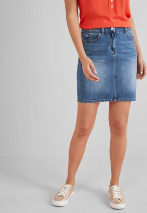 Femme portant une jupe en jean bleue, un haut sans manches orange et des baskets beiges, debout devant un fond gris clair uni.