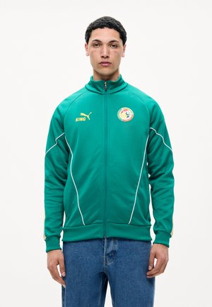 Jonge man draagt een groene Puma jas met ritssluiting met het logo van het Senegalese voetbalteam en de tekst "KING", gecombineerd met een blauwe spijkerbroek, kijkt naar voren.
