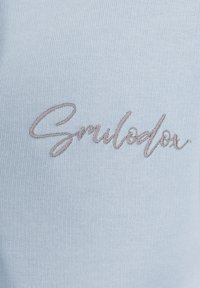 Un tessuto azzurro chiaro presenta il testo ricamato "Smilodox" in un carattere corsivo, mostrando una texture liscia e un design minimalista.