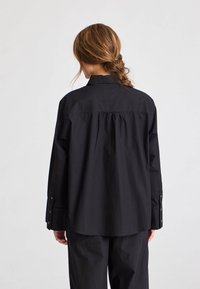 Cămașă neagră cu m�ânecă lungă, cu croială lejeră, dintr-un material texturat, cu mansete cu nasturi și detaliu de yoke pe spate, asortată cu pantaloni potriviți.