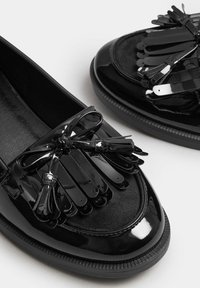 Svarta lackskinnsloafers med dekorativ frans och rosett, slät textur, rund tå och flat gummisula.