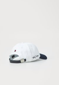 White cotton cap with navy brim, featuring embroidered signature and small logo patch on the back. Adjustable strap in navy with silver hardware.