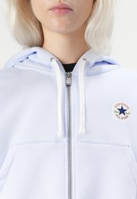 Sudadera azul claro con cremallera hecha de algodón. Presenta cordones blancos, un bolsillo frontal de parche y un logo redondo con una estrella en el pecho.