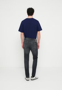 Mies seisoo selin kameraan, päällään tummansininen t-paita, haalistuneet mustat farkut, mustat sukat ja valkoiset tennarit valkoista taustaa vasten.