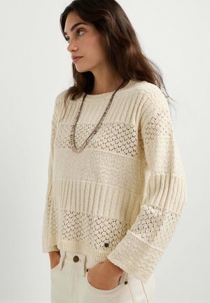 Femme portant un pull en maille crème avec des motifs texturés et en dentelle, un long collier et un pantalon crème, debout les mains dans les poches.