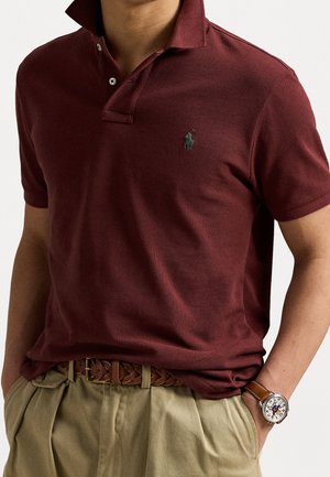 Person iført en mørkerød poloshirt med et grønt logo, beige plisserede bukser, et brunt flettet bælte og et armbåndsur med en brun læderrem.