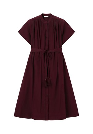 Femme en robe midi bordeaux boutonnée avec manches retroussées, portant des sandales beiges à talons avec des nœuds décoratifs, debout, pensive.