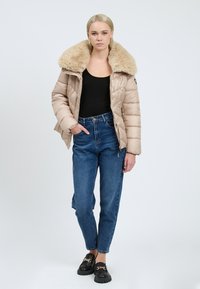 Puffet beige jakke med en stor krave af kunstpels, parret med højtaljede blå jeans og sorte loafers med gyldne kæde detaljer.
