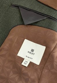 Brązowa tkanina z teksturowanym, geometrycznym wzorem, z białą metką oznaczoną "NEXT SLIM FIT" oraz rozmiarem "40R". Tło jest ciemnozielone.