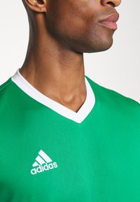 Grønn atletisk trøye med hvit V-halskant. Har Adidas-logoen i hvitt på venstre side. Glatt, pustende stoff.