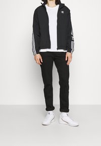 Svart träningsjacka med vita ränder, dragkedja, logotyp på bröstet, tillsammans med en vit t-shirt, svarta byxor och vita sneakers.